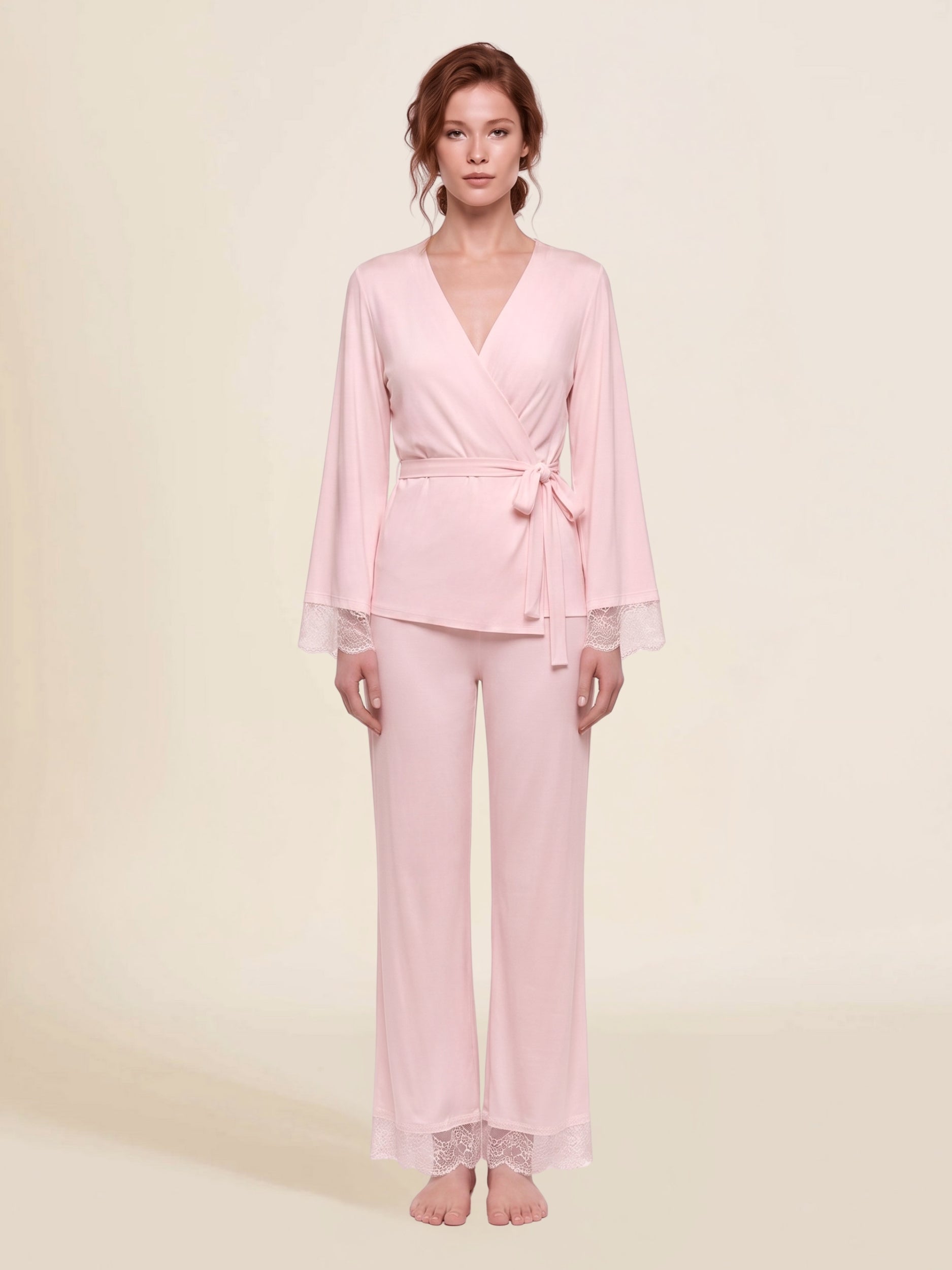 A woman in a light pink micro modal and lace two-piece luxury pajama set consisting of a long-sleeve wrap top with bell sleeves and lace cuffs and matching palazzo pants with lace hem.