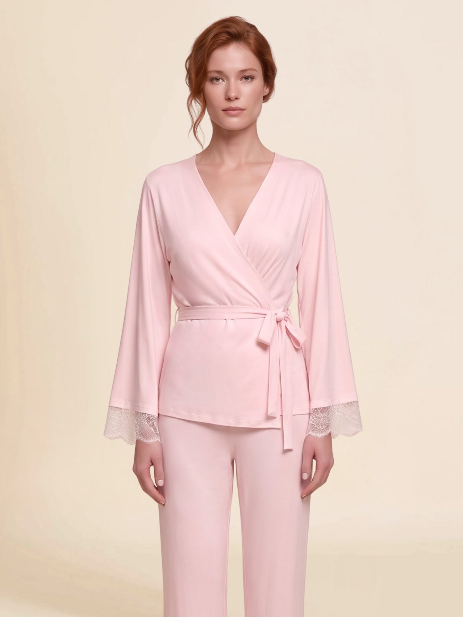 A woman modeling a light pink micro modal and lace wrap-style top with long bell sleeves, wide lace trim at the cuffs, and a matching tie belt, paired with matching light pink palazzo pants with lace cuffs.